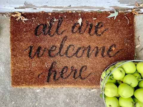 Doormat reading "All are welcome here," a bushel of green apples is in the bottom right corner